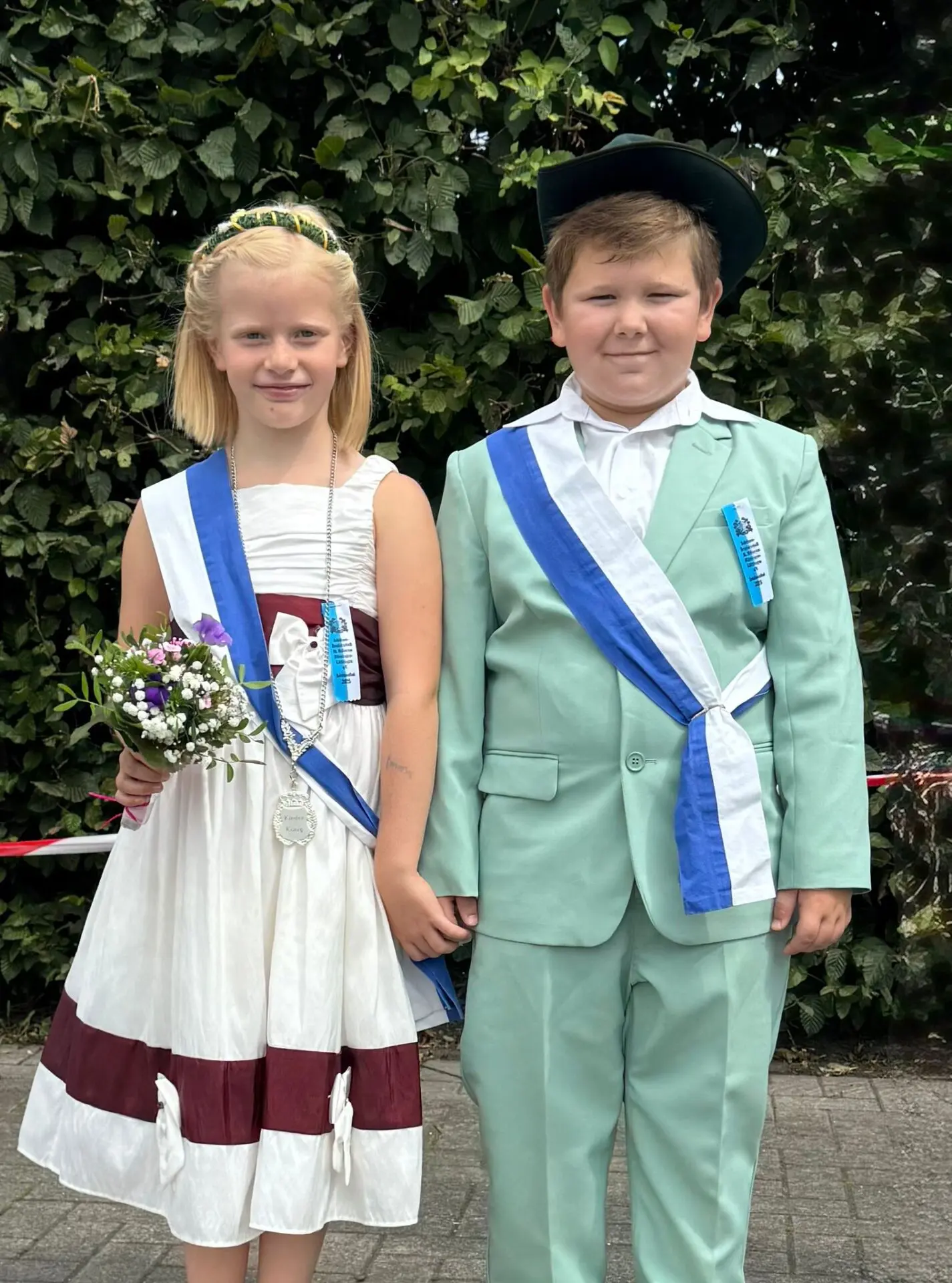 Ein Junge und ein Mädchen in festlicher Kleidung stehen nebeneinander, beide mit Schärpen und einer Blumenstrauß.
